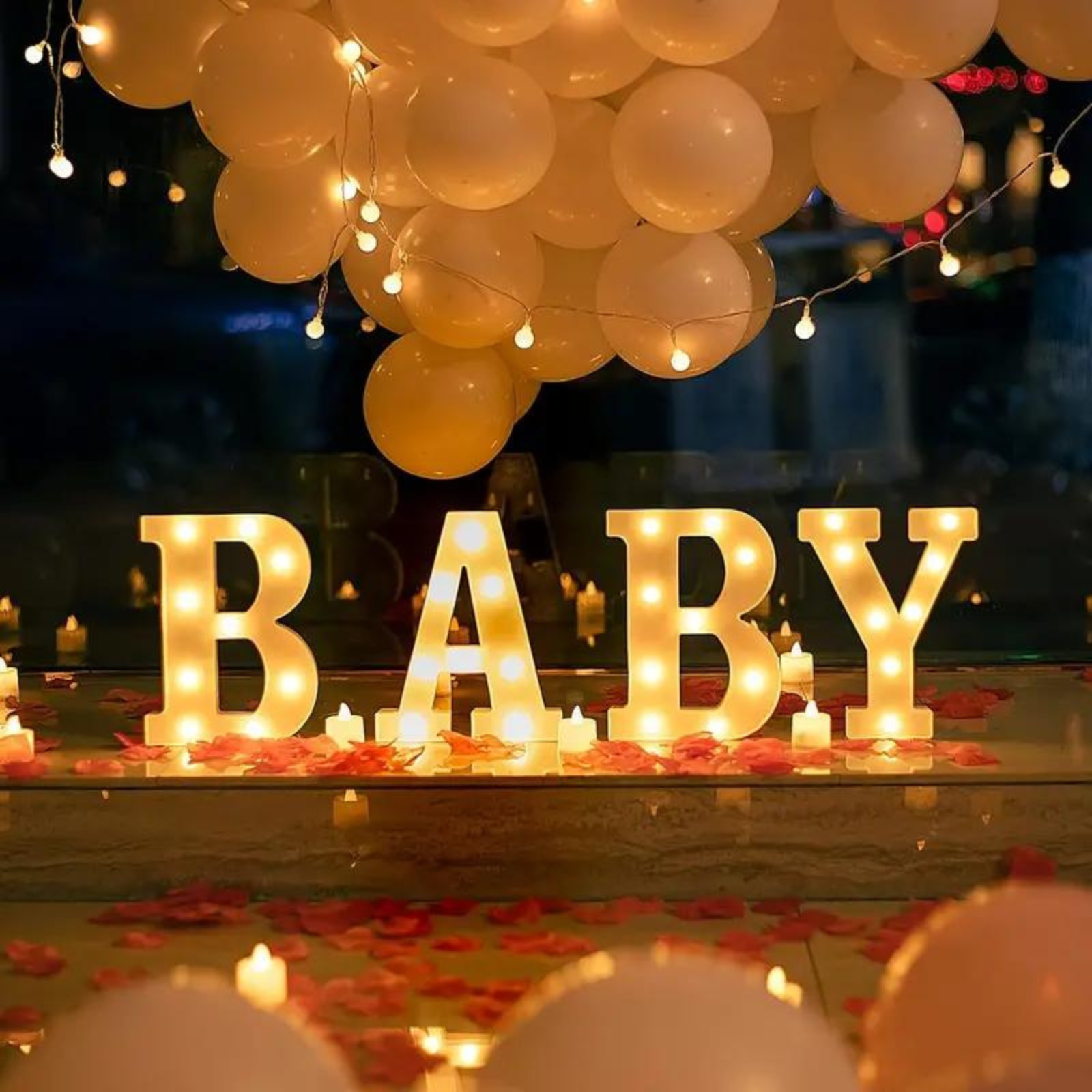 Illuminated ‘BABY’ letters surrounded by candles, rose petals, and balloons in a warm, elegant setup.