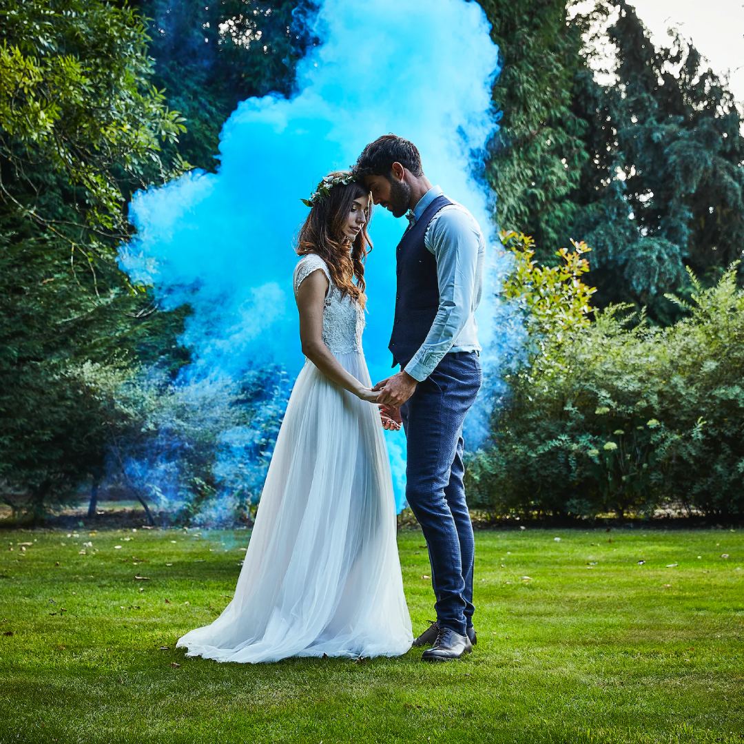 Couple standing close together with a blue smoke cloud behind them, wearing formal attire, as part of a gender reveal celebration